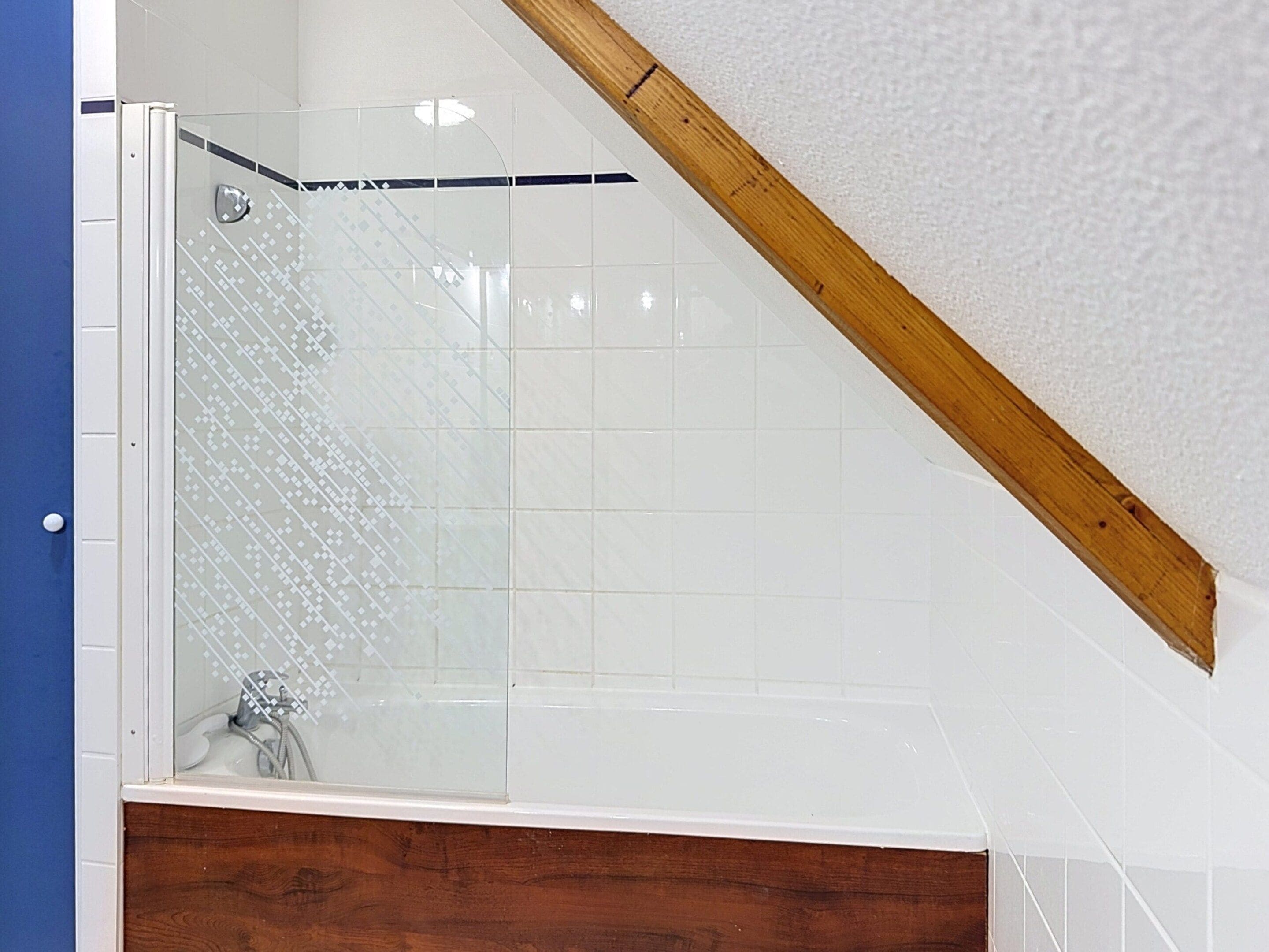 A shower with a glass door, a wooden ceiling beam, and a white tiled wall.