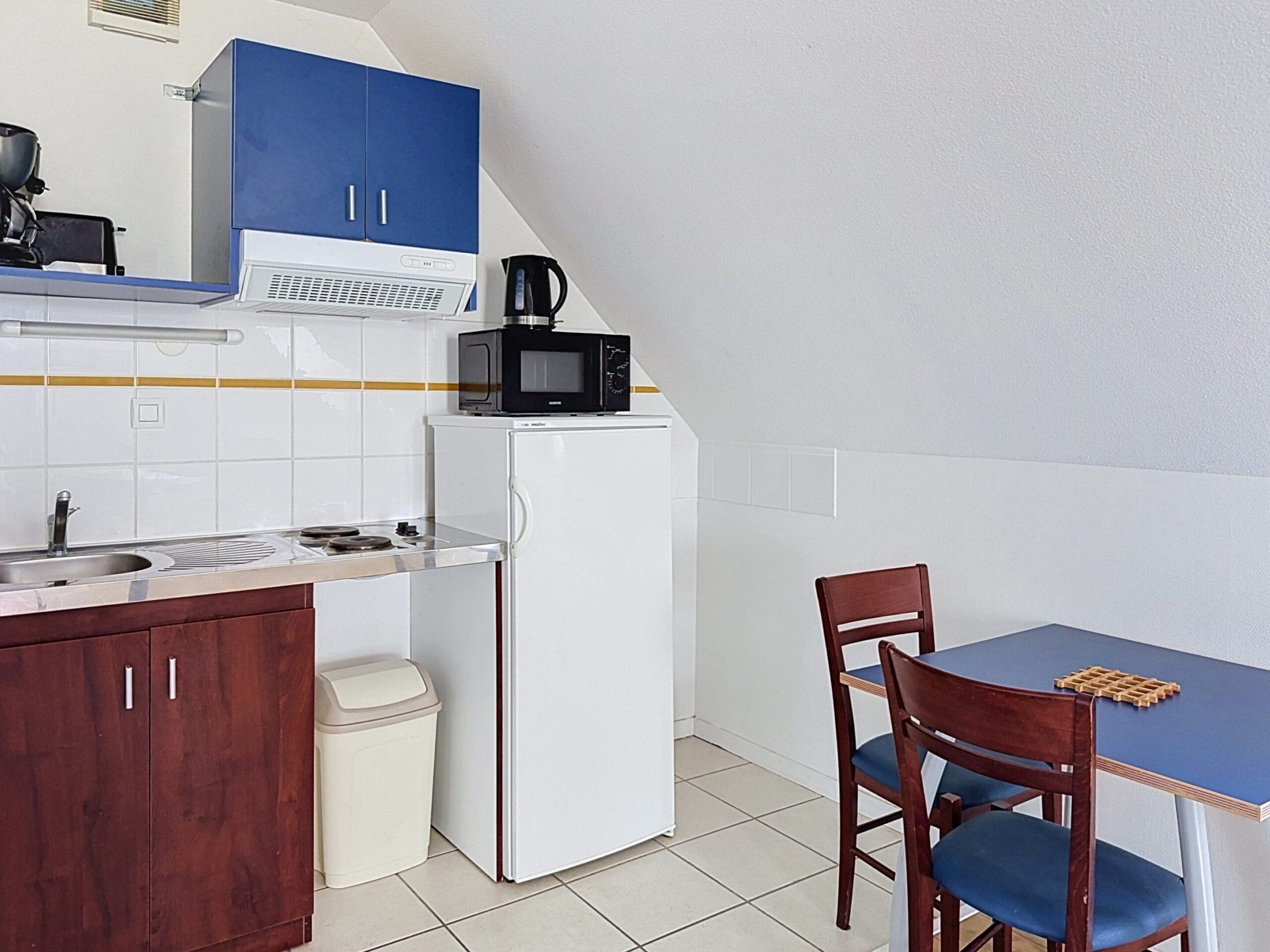 Een kleine keuken met een witte koelkast, een magnetron, een spoelbak en een eettafel met stoelen.