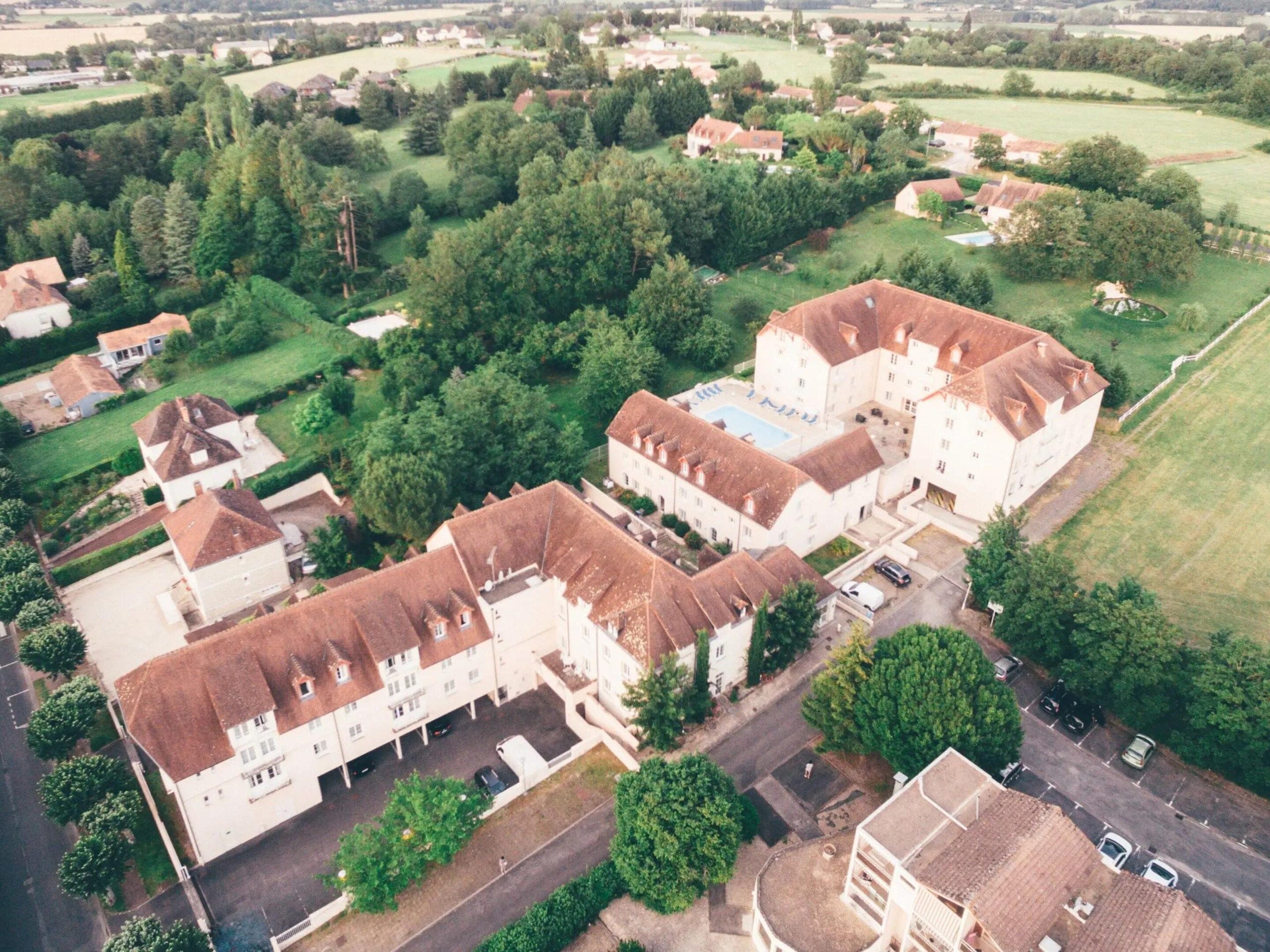 An aerial view of a residential area with multiple houses, a large building with a swimming pool, and surrounding greenery.