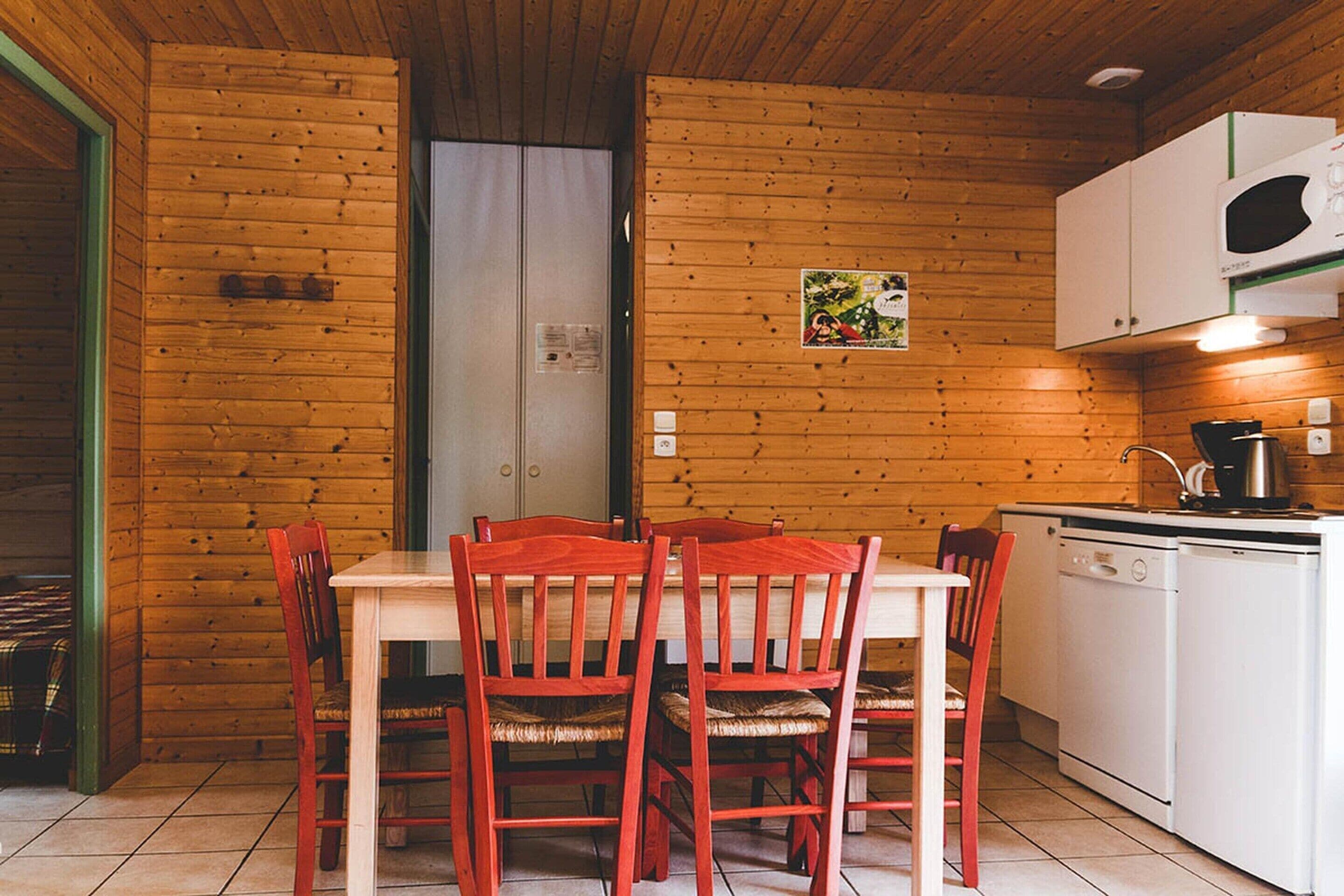 A wooden interior with a dining table, chairs, a kitchen area, and a door.