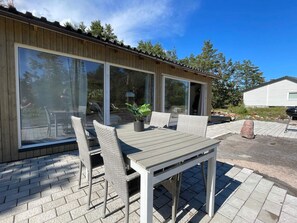 A modern outdoor patio with a wooden table and wicker chairs, large sliding glass doors, and a view of the surrounding forest. - 4 Star Holiday Home in Oskarshamn (Oskarshamn)