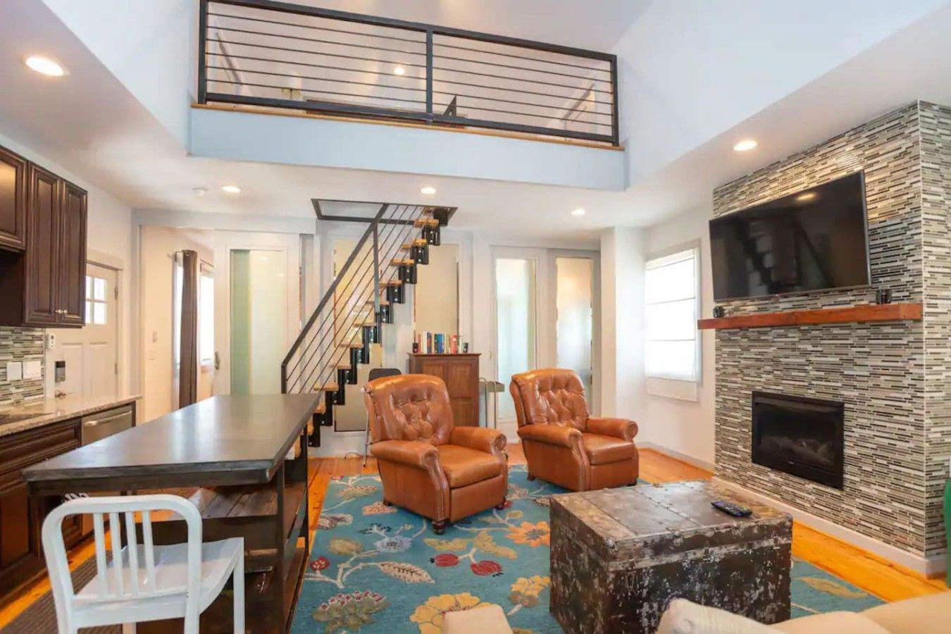 A spacious living area with a high ceiling, a fireplace, a staircase, and a kitchen area.