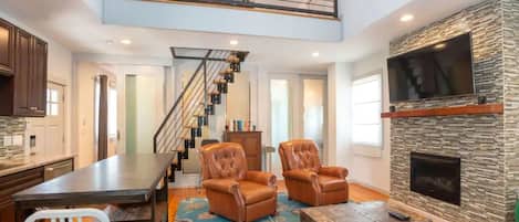 A spacious living area with a high ceiling, a fireplace, a staircase, and a kitchen area.