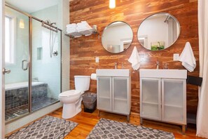 Ein Badezimmer mit Duschkabine aus Glas, zwei Waschbecken mit gefrosteten Schränken, einer Toilette und zwei runden Spiegeln.