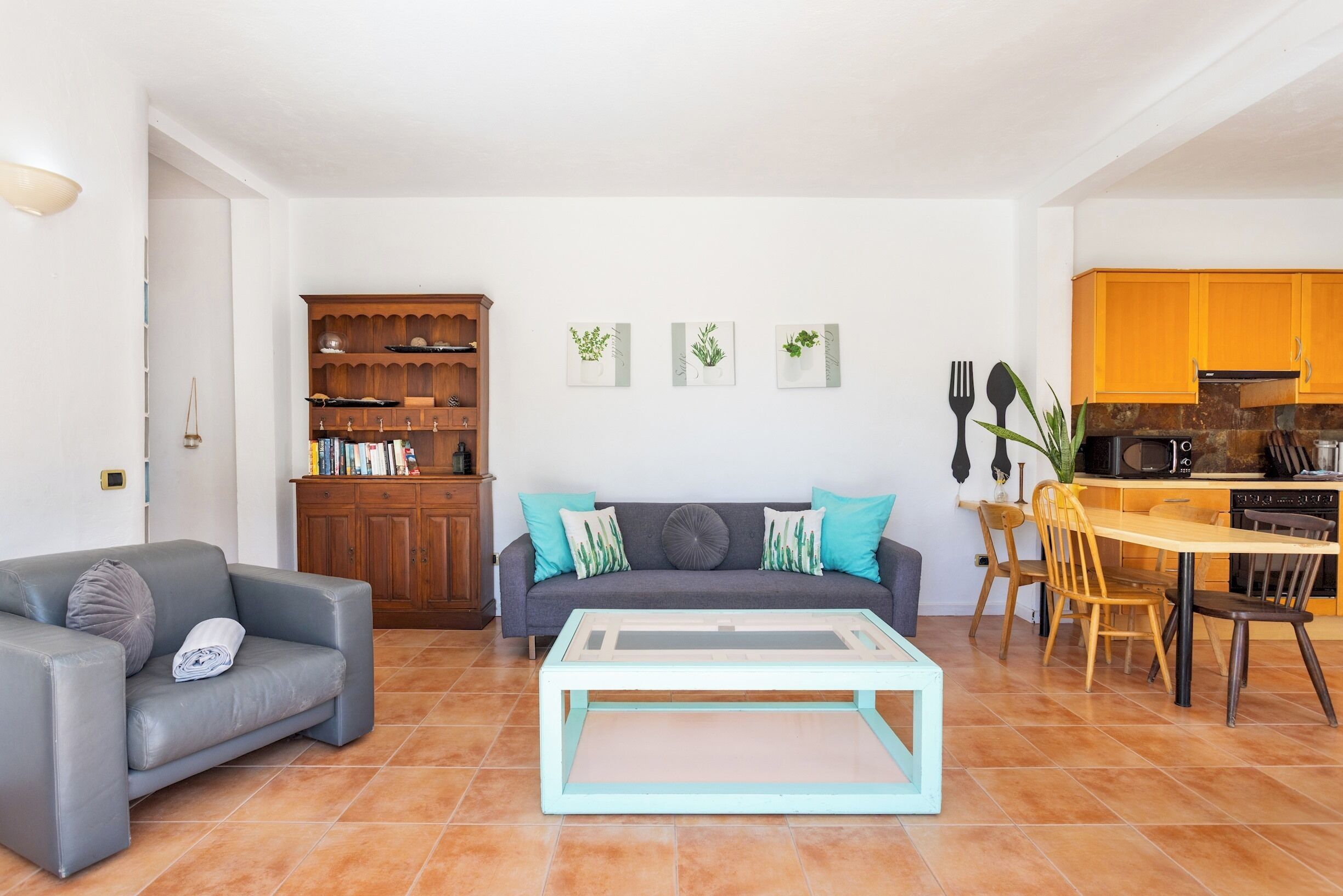 A living room with a sofa, a coffee table, a bookshelf, and a kitchen area with wooden cabinets and a dining table.