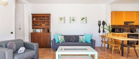 A living room with a sofa, a coffee table, a bookshelf, and a kitchen area with wooden cabinets and a dining table.