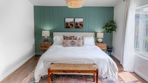 Een slaapkamer met een bed, twee nachtkastjes, een bankje, een hanglamp en een raam met gordijnen.