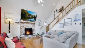 Ein Wohnzimmer mit Kamin aus Stein, einem grauen Sofa, einem roten Couchgarnitur mit Muster und einer Holztreppe, sowie einem Gemälde eines Sees.