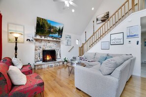Ein Wohnzimmer mit Kamin aus Stein, einem grauen Sofa, einem roten Couchgarnitur mit Muster und einer Holztreppe, sowie einem Gemälde eines Sees.