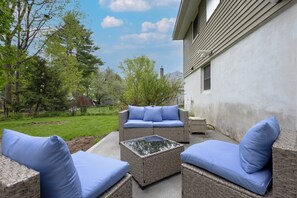 Ein Sitzbereich im Freien mit Korbmöbeln und blauen Kissen, einem Glastisch und Blick auf ein Haus und Bäume.