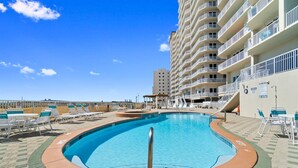 A swimming pool with a curved edge, surrounded by lounge chairs and a building with multiple balconies. - Beachfront Condo | Steps to Sand | 3 Pools (Navarre)