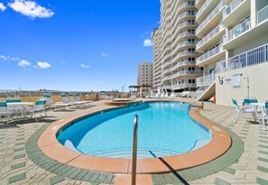 Une piscine à bordures courbes, entourée de chaises longues et bordée d’un bâtiment doté de nombreux balcons.