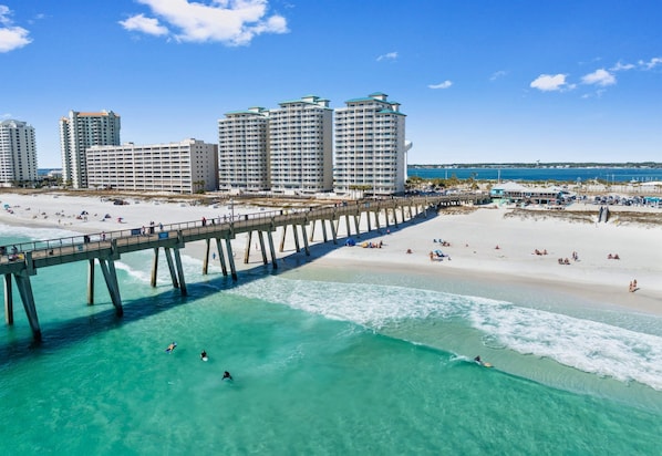 A beach with a pier, clear turquoise water, and high-rise buildings in the background.
