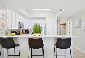 A modern kitchen with white cabinets, a central island with bar stools, a microwave, and a potted plant. - Beachfront Condo | Steps to Sand | 3 Pools (Navarre)