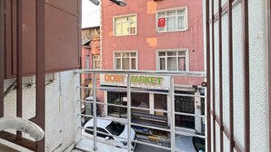 Een balkon met uitzicht op een gebouw met een 'DOST MARKET' bord, een witte auto geparkeerd daaronder, en een metalen hekwerk.