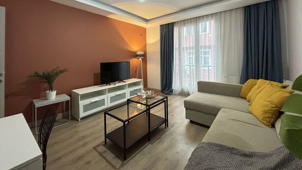 Een moderne woonkamer met een flatscreen-tv, een salontafel, een bank met kussens, een lamp en een raam met gordijnen.