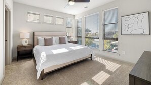 A bedroom with a bed, nightstands, a ceiling fan, large windows with blinds, and a view of the outside. - Landing Furnished Apartments - Port St. Lucie (Port Saint Lucie)