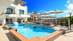 Ein moderner Poolbereich im Freien mit einem kühlen, blauen Pool, Liegestühlen, Sonnenschirmen und einem mehrstöckigen Gebäude mit Balkonen.