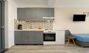 A compact kitchen with grey cabinets, a white stove, and a small dining area with a chair and a bed.