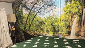 Een slaapkamer met een groot muurdecoratie van een bos en waterval, een bed met een groene deken en een houten vloer.
