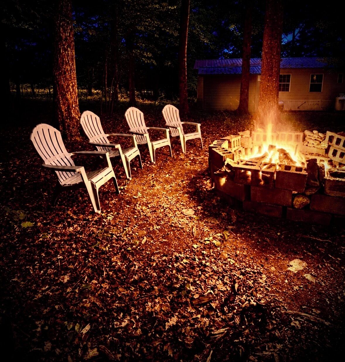 A campfire with chairs around it, a brick fire pit, and a house in the background.