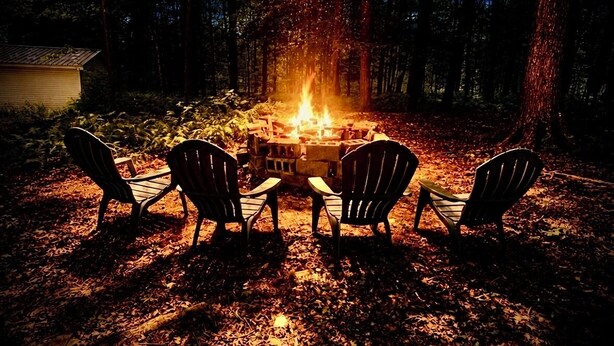 A campfire with four Adirondack chairs arranged around it in a forest setting.