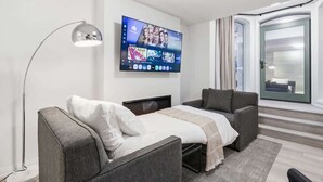 Quarto de hotel moderno com uma cama, duas poltronas, uma televisão fixada na parede e uma porta de vidro deslizante que dá para uma varanda.