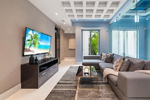 A modern living room with a flat-screen TV, a dark wooden entertainment unit, a grey sofa, and a glass partition wall.
