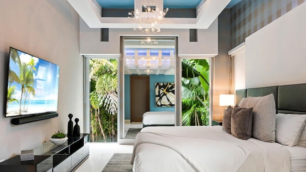 A modern bedroom with a large bed, a TV on the wall, a chandelier, and a view of greenery through the door. - Luxe Grove Villa with private pool (Miami)