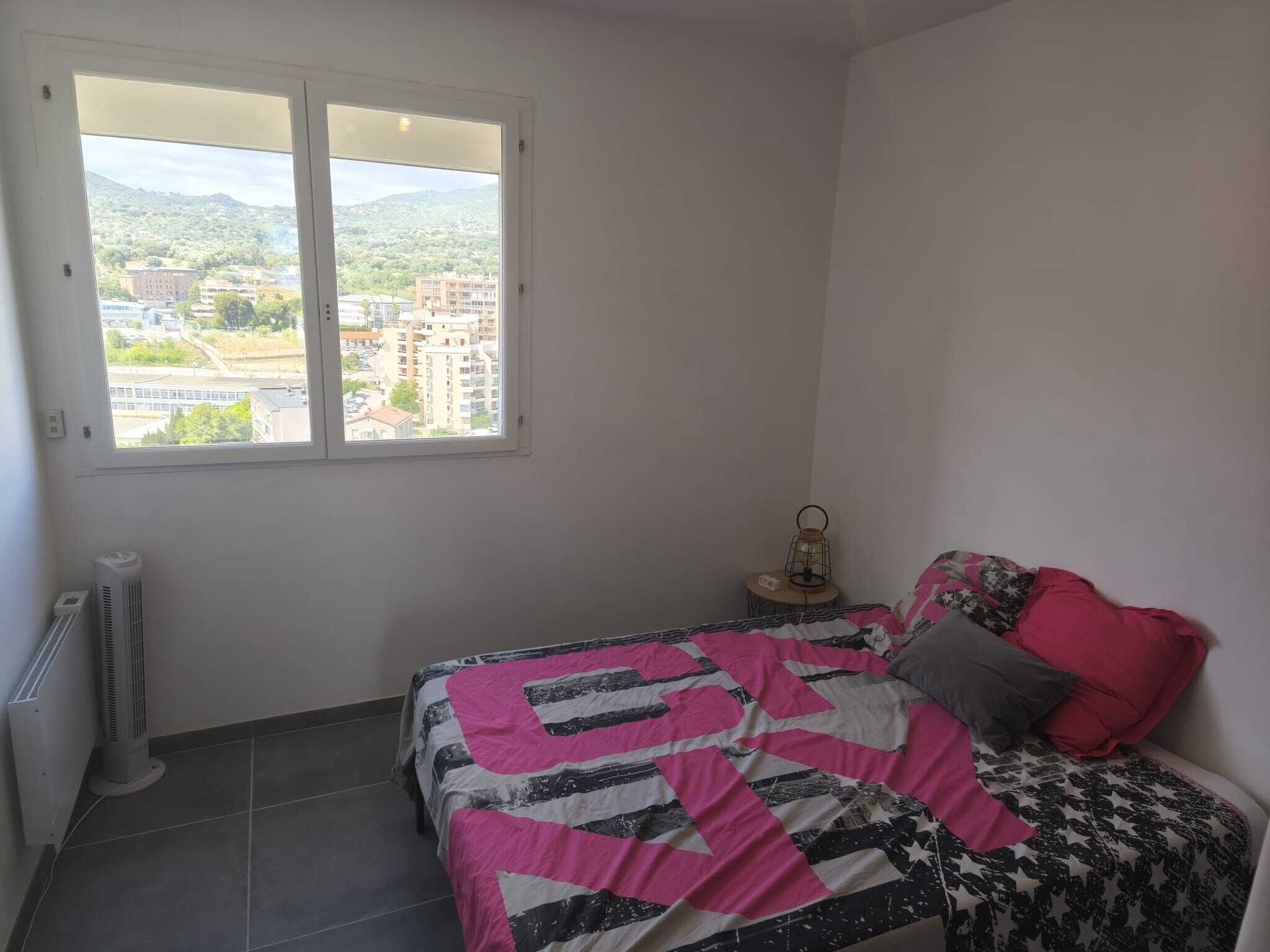 Quarto com cama, janela com vista para edifícios, radiador e uma pequena mesa com um candeeiro.