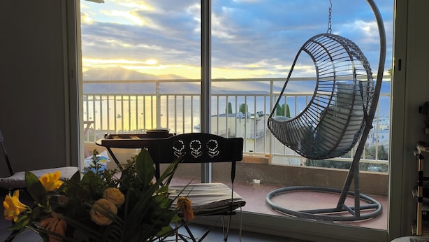 Un balcon agrémenté d’un fauteuil suspendu, une table ornée d’un vase de fleurs et une vue sur la mer et le paysage urbain.