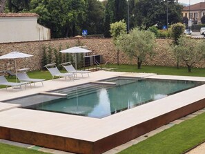 A rectangular swimming pool with a stone wall and a grassy area with lounge chairs and umbrellas. - Elegant Suite With Shared Pool (Adro)