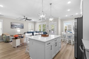 A modern kitchen with white cabinetry, a central island, pendant lighting, and a dining area with white chairs and a table. - Ocean Lakes Site T37 - Beachside Blessings (Myrtle Beach)