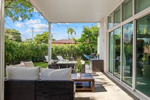 Une terrasse couverte, aménagée avec du mobilier d’extérieur et une table à manger, offrant une vue sur un jardin luxuriant et les bâtiments voisins.