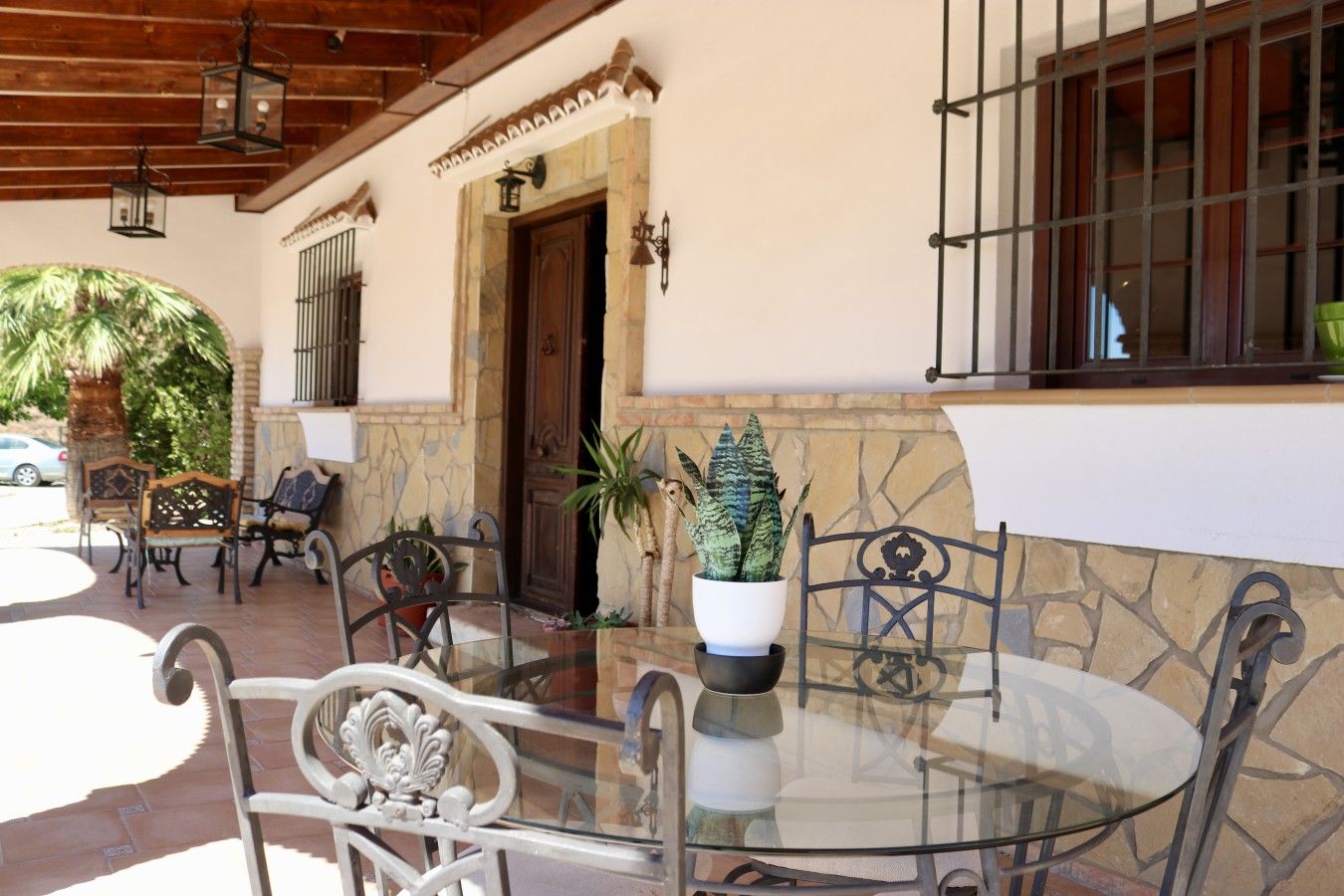 An outdoor seating area with a glass table and metal chairs, a potted plant, and a stone wall.