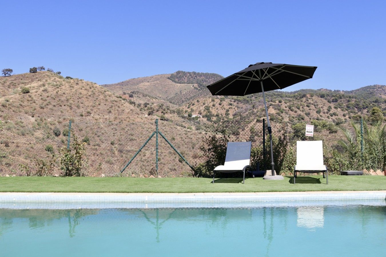 A swimming pool with two lounge chairs and an umbrella, surrounded by a fence and hills in the background.