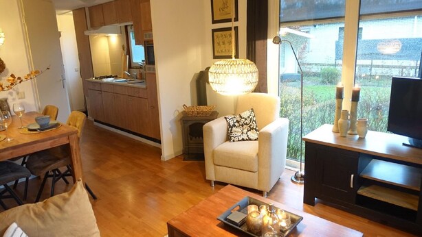 A modern living room with a sofa, coffee table, and a TV stand. There's a dining area with a table and chairs. The room has a large window with a view of greenery outside. - Vechtdal Chalet de Eik (Rheezerveen)