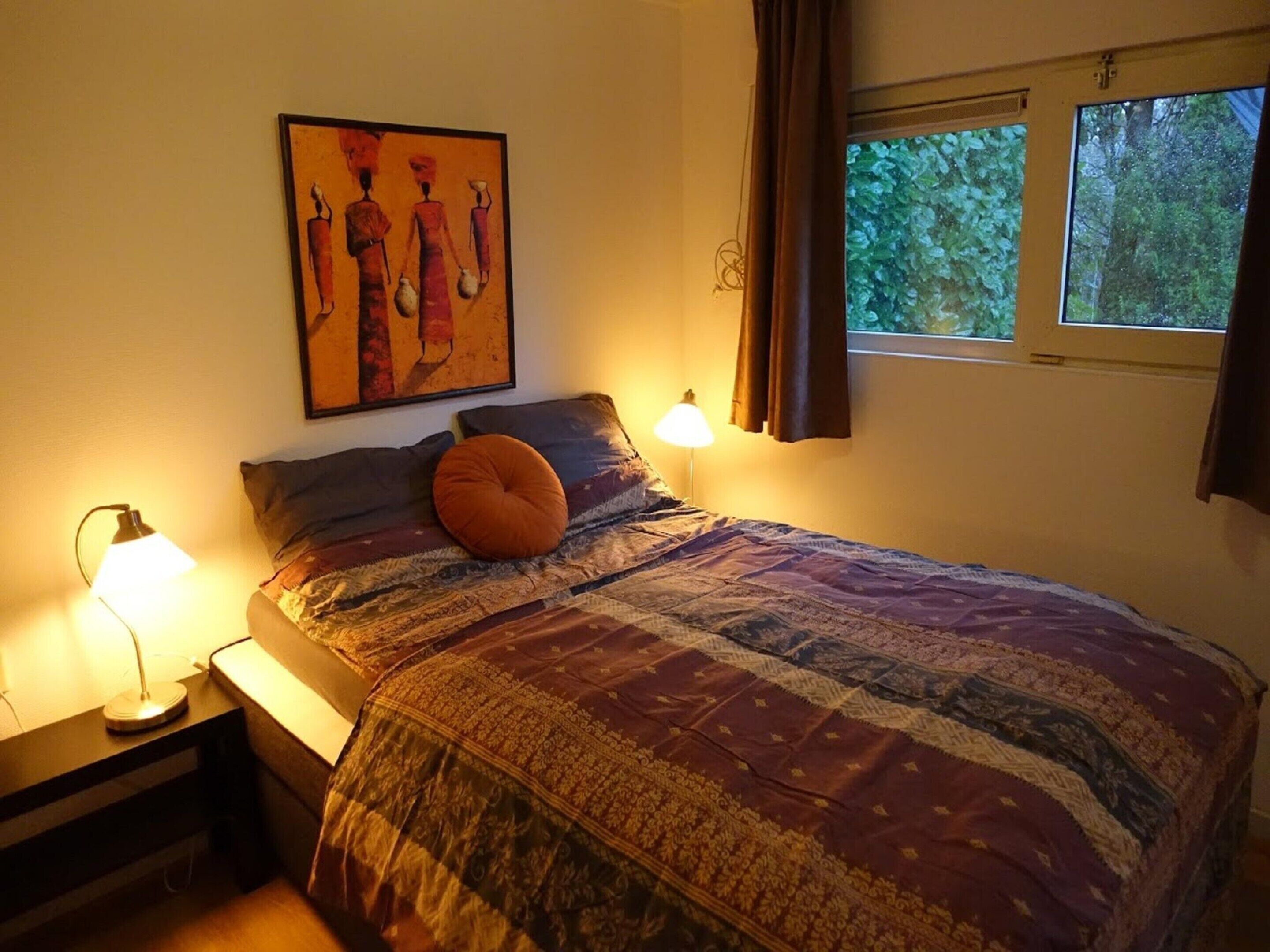 A bedroom with a bed, a nightstand, a lamp, a painting, and a window with curtains.