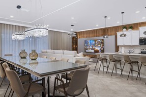 Cozinha moderna com armários brancos, área de jantar com mesa de vidro e sala de estar com sofá e cortinas.