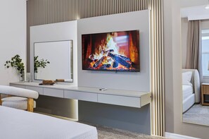 Ein modernes Hotelzimmer mit einem an der Wand befestigten Flachbildfernseher, einem Nachttisch mit Buch, Pflanze und einem weißen Bett.