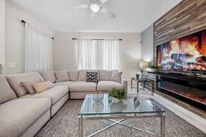 Ein Wohnzimmer mit einem Ecksofa, einem Glastisch, einem Kamin und einem Fernseher.