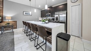 Una cocina moderna con una isla central, taburetes de bar, electrodomésticos de acero inoxidable, iluminación colgante y un reloj en la pared.