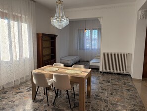 Un comedor con una mesa de madera, cuatro sillas, una lámpara de araña, una estantería, un radiador y una ventana con cortinas.