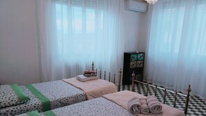 Une chambre d’hôtel avec deux lits, un sol à carreaux, une armoire et une fenêtre avec des rideaux légers.