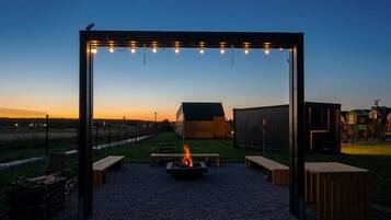 Un espace extérieur aménagé avec un foyer, des bancs et une pergola illuminée de guirlandes lumineuses.
