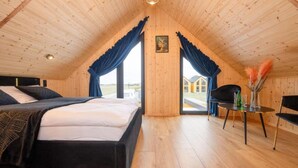 A high-ceilinged room with a bed, a table, and a chair, with a view of buildings outside.