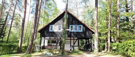 A cabin with a gabled roof, surrounded by tall trees, with a dirt path leading to the entrance.