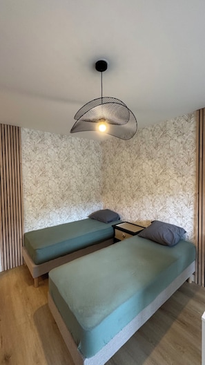 A room with two beds, a patterned wallpaper, a hanging light fixture, and wooden paneling. - Studio Bagnole de l'Orne Quiet and Bright (Bagnoles de l'Orne Normandie)