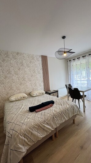 A bedroom with a large bed, a bedside table, a chair, a window with curtains, and a ceiling light fixture. - Studio Bagnole de l'Orne Quiet and Bright (Bagnoles de l'Orne Normandie)