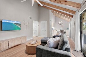 Ein modernes Wohnzimmer mit Fernseher, einem grauen Sofa, einem Holztisch und Blick auf den Strand durch das Fenster.